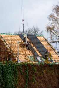 Workers secure a waterproof covering over an open roof section beside stacked lumber and renovation tools.